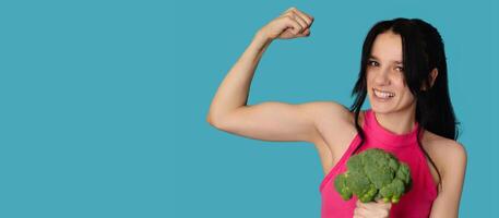 A smiling white girl with fresh broccoli in one hand and showing off her biceps in the other. Panoramic banner, free space. photo