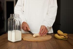 Close up of young woman with blender chopping banana for milkshake at home photo