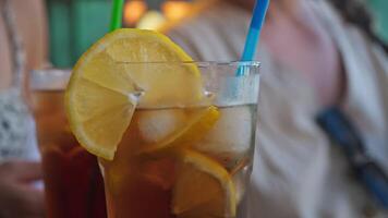 Close-up of a slice of lemon, perched on rim of a glass, garnishing a chilled cocktail with ice cubes on a table in a bar. In the background, people are conversing. Slow motion. video