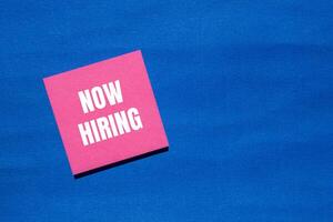 Now hiring message written on pink paper with blue background. Conceptual now hiring symbol. Copy space. photo