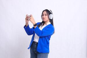 beautiful office girl wearing blue formal suit standing slanted right pouting to camera both hands playing game on smartphone mobile phone and headphone isolated white background. for entertainment photo