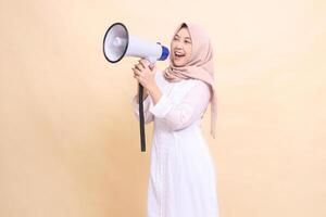 woman asian young wearing hijab slanted to the right candidly shouts cheerfully holding a loudspeaker megaphone with both hands. Technology, broadcast and promotion concept photo
