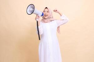 girl asian muslim wearing a hijab slanted to the right, candid, shouting, smiling, holding a loudspeaker megaphone and clenching her fists upwards. Technology, broadcast and promotion concept photo