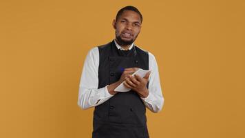 Professional caterer writing clients orders on notepad for the bill, takes notes of desired food against yellow background. Waiter with apron, notebook service for customer satisfaction. Camera B. photo