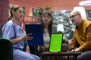 Medical assistant visiting elderly couple for checkup at their home, using isolated display and doing a consultation to give treatment and vitamins for recovery. Nurse talking to senior patients. photo