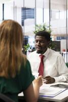 Headhunter tasked with hiring best talent to fill open company position, interviewing man. Employer hired to assess skills and qualifications of african american candidate, doing meeting in office photo