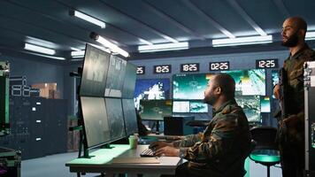 Military agent working in control room, analyzing aerial reconnaissance data and talking with colleague. Intelligence corps agent checking screens, discussing with coworker, camera B photo
