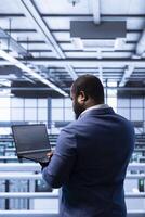 Data center manager ensuring safety and security of database using notebook. African american man in server farm preventing unauthorized access and suspicious activities posing risks for business photo