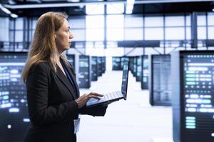 IT staff member walking in data center, using laptop to configure network infrastructure such as routers, switches and firewalls. Woman working in server room looking for technical issues photo