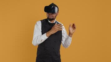 Restaurant waiter uses virtual reality headset with artificial intelligence, having fun with technology and vr glasses against yellow background. Employee with apron and uniform. Camera B. photo