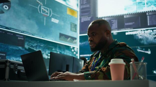 IT analyst in a high tech base of operations handling surveillance activity and ensuring security protocols. Military command post crew collecting real time data to achieve objectives. Camera B. photo