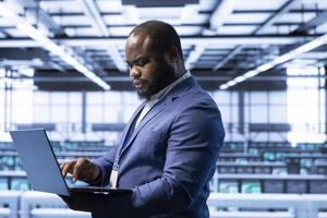 Computer scientist using laptop in data center with advanced tech, ensuring cybersecurity and efficient network management. African american expert in server hub optimizing data center with device photo