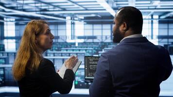 Teamworking programmers in data center overseeing server operations using laptop and tablet. Coworkers ensuring optimal performance and data integrity in server room using devices, camera A photo
