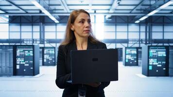 Software developer walking in data center, overseeing server operations using laptop. Woman ensuring optimal performance and data integrity in server farm using notebook, camera B photo