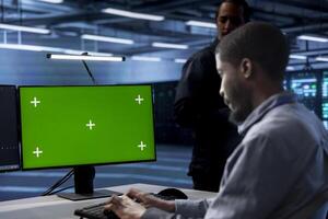 Focused software developer working on computer with green screen monitor in modern data center. IT professional doing IT infrastructure management on server racks in high tech environment photo