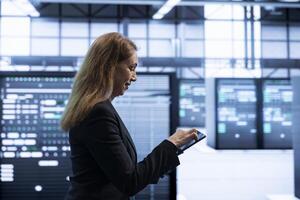 Programmer using tablet in data center with advanced tech, ensuring cybersecurity and efficient network management. IT specialist in server hub optimizing data center infrastructure with device photo