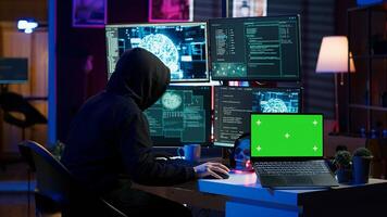 Hacker in hidden place using green screen laptop to deploy malware on unsecured devices, stealing sensitive data from unaware users online and selling it on black market, camera A photo