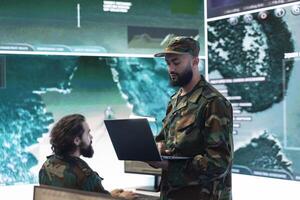 Staff sergeant briefing his military crew with details of a new secret service mission, operating high tech in command post. Army leader monitors radar CCTV footage with a satellite world map. photo