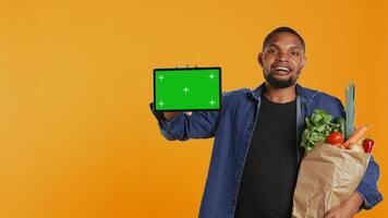 African american guy showing a tablet with greenscreen layout, going on a grocery shopping spree and supporting local farming. Young man advertises an isolated screen and healthy nutrition. Camera B. photo
