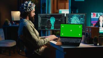 Engineer with EEG headset on programming brain transfer into computer virtual world next to green screen laptop. Transhumanist using neuroscience to gain digital soul, mockup device, camera A photo