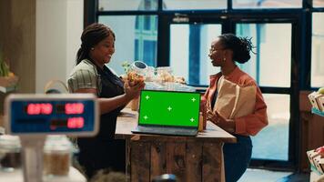 Seller presenting her merchandise next to greenscreen on pc, customer visiting local farmers market to buy bulk products. Merchant recommending grains and pasta, laptop runs isolated chromakey. photo