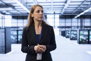 IT staff member walking between server rows in data center, doing yearly infrastructure inspection. Woman in high tech server room facility monitoring supercomputers equipment photo