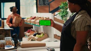 African american vendor using greenscreen template at counter, presenting chromakey display on smartphone. Woman seller holding phone with isolated mockup layout and copyspace. photo