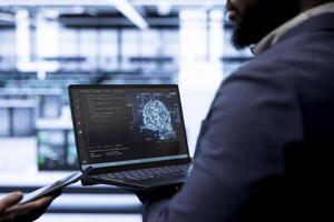 Engineer using AI software on laptop, programming in data center, updating neural networks. Close up shot of IT professional in modern server hub startup upgrading artificial intelligence systems photo