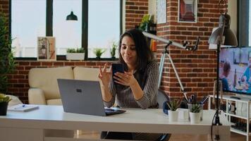 Teleworker checking her phone notifications at home in front of laptop, taking break from work. Woman in cozy apartment office taking pause from job tasks to read messages on smartphone, camera A photo