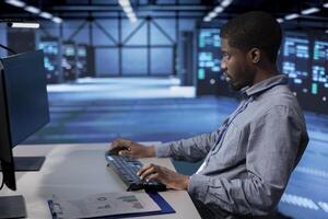 African american man typing on PC in server farm, ensuring data remains shielded from potential threats. IT expert coding at computer desk, safeguarding data center hardware against malicious access photo