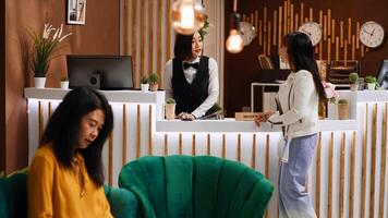 Asian client arriving at reception desk with trolley bags, staff asking tourist about her room reservation for check in process. Hotel concierge greeting guest at holiday resort, tourism concept. photo
