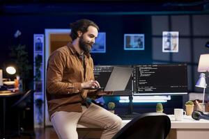 Smiling IT expert doing software quality assurance, reading source code on laptop before implementing it. Happy man sitting on desk inspecting coding on notebook, looking to fix potential issues photo