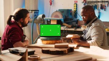 Joyous cabinetmaker looks over blueprints schematics on isolated screen laptop in CAD software with apprentice. Smiling artisan and colleague designing custom 3D furniture on mockup notebook, camera A photo
