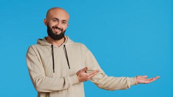 Cheerful man creating ad in front of studio camera, pointing to left or right sides over blue background. Middle eastern person showing commercial or advertisement for marketing campaign. Camera 2. photo