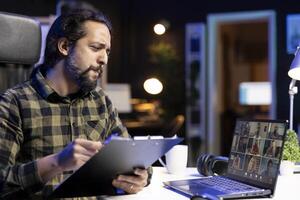 Young adult working from home, going over his notes while on a call with a variety of people. Male freelancer using his laptop and holding his clipboard during a virtual meeting. photo