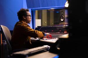 African american sound designer operates audio console and software on pc, moving faders and pressing switchers to edit tracks. Audio expert works in professional studio control room. photo