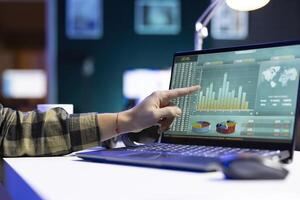 An individual pointing at a screen that shows numbers and financial charts related to stock trading. At a desk, digital laptop is displaying graphs and values for forex trends. photo