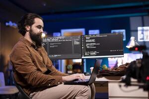 Programmer doing software debugging using laptop, checking errors in lines of code displayed on computer monitors. Man working from home fixing coding mistakes using notebook device photo