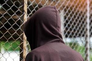male teenager wearing a brown hoodie stood facing a steel mesh fence. Both hands cling to the fence, showing the back. photo
