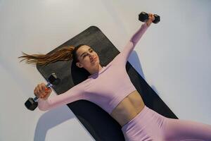 Top view of strong woman lying on yoga mat and doing exercises for hands muscles with dumbbells photo