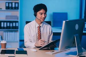 Happy smiling Asian man work at call center service desk consultant with teammates at night, customer service executive with microphone headset use computer for supporting in late night office. photo