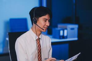 Happy smiling Asian man work at call center service desk consultant with teammates at night, customer service executive with microphone headset use computer for supporting in late night office. photo