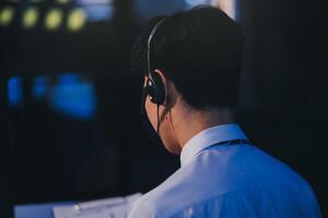 Happy smiling Asian man work at call center service desk consultant with teammates at night, customer service executive with microphone headset use computer for supporting in late night office. photo