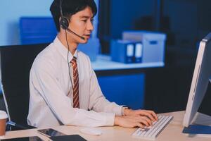 Happy smiling Asian man work at call center service desk consultant with teammates at night, customer service executive with microphone headset use computer for supporting in late night office. photo