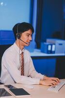Happy smiling Asian man work at call center service desk consultant with teammates at night, customer service executive with microphone headset use computer for supporting in late night office. photo