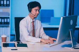 Happy smiling Asian man work at call center service desk consultant with teammates at night, customer service executive with microphone headset use computer for supporting in late night office. photo