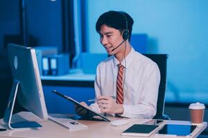 Happy smiling Asian man work at call center service desk consultant with teammates at night, customer service executive with microphone headset use computer for supporting in late night office. photo