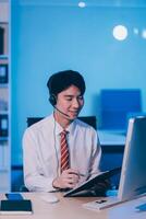 Happy smiling Asian man work at call center service desk consultant with teammates at night, customer service executive with microphone headset use computer for supporting in late night office. photo