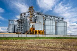 large grain elevator with multiple cylindrical metal silos, structural elements, and a complex system of pipes. It stands against a blue sky, with a field in the foreground after harvest photo