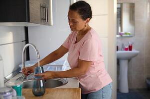Woman in casual clothes filling a glass at kitchen sink in a cozy home setting, illustrating daily routine and household chores with a sense of tranquility photo
